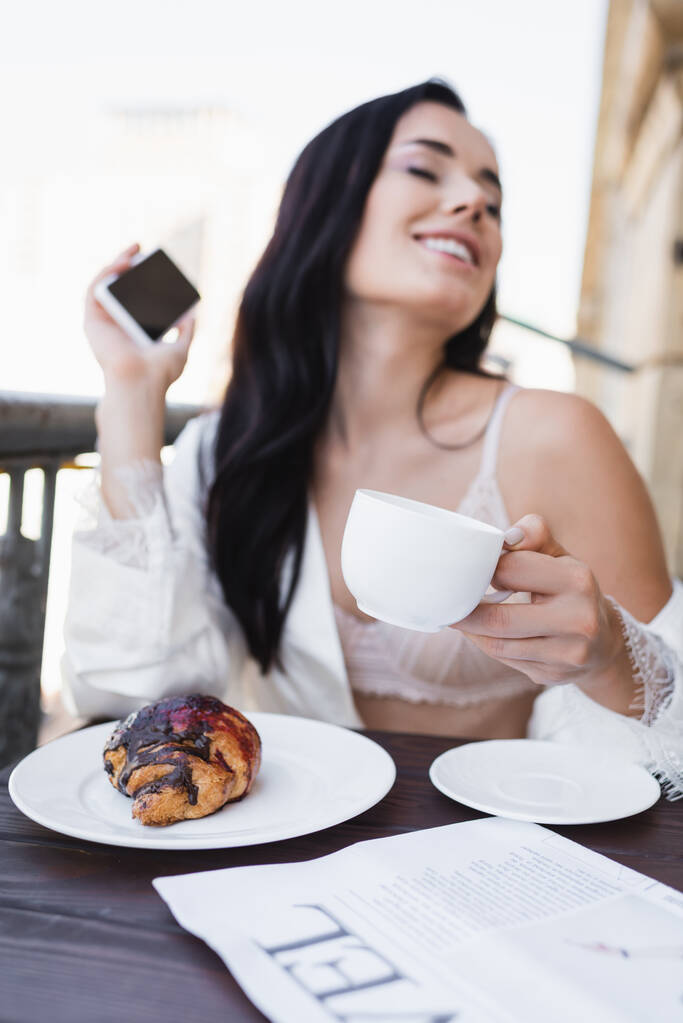 kaunis brunette nainen valkoinen kaapu tilalla älypuhelin ja ottaa aamiaisen parvekkeella - Valokuva, kuva