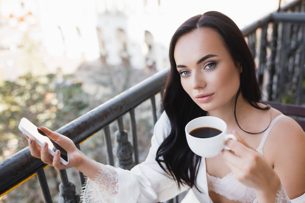 schöne brünette Frau im weißen Gewand, die Kaffee auf dem Balkon trinkt und ihr Smartphone hält - Foto, Bild