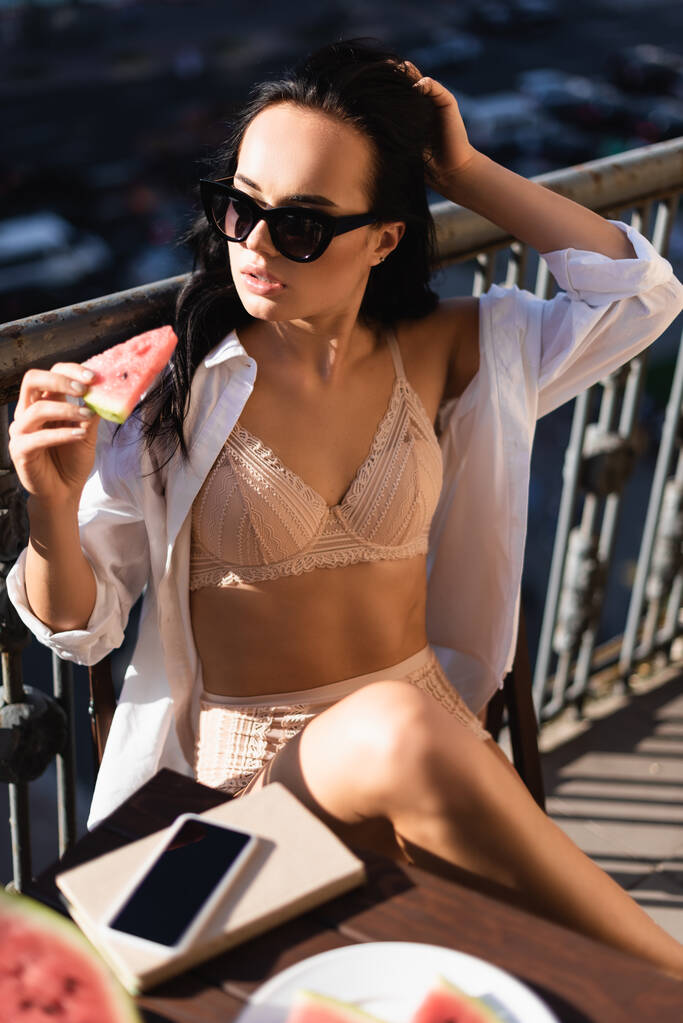 brunette woman in beige underwear and white shirt eating watermelon on balcony - Photo, Image
