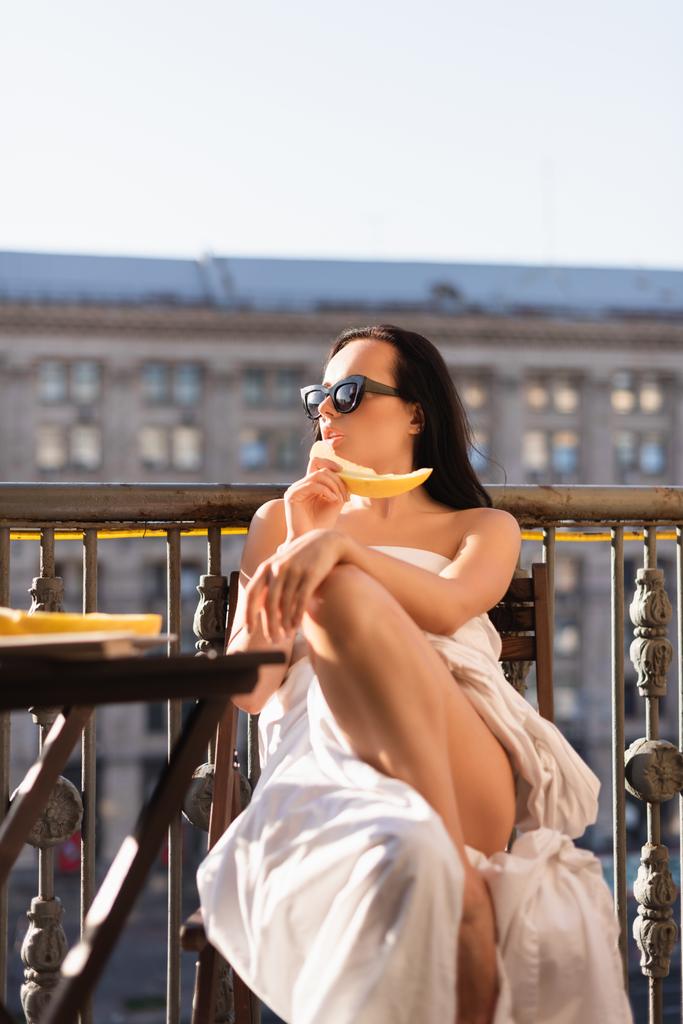 sexy brunette vrouw in zonnebril en bedekt met wit laken eten meloen op balkon - Foto, afbeelding