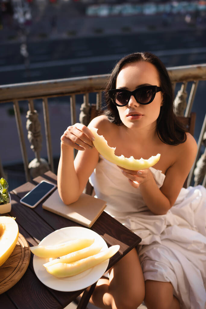 σέξι μελαχρινή γυναίκα με γυαλιά ηλίου και καλυμμένη με λευκό σεντόνι τρώγοντας πεπόνι στο μπαλκόνι - Φωτογραφία, εικόνα