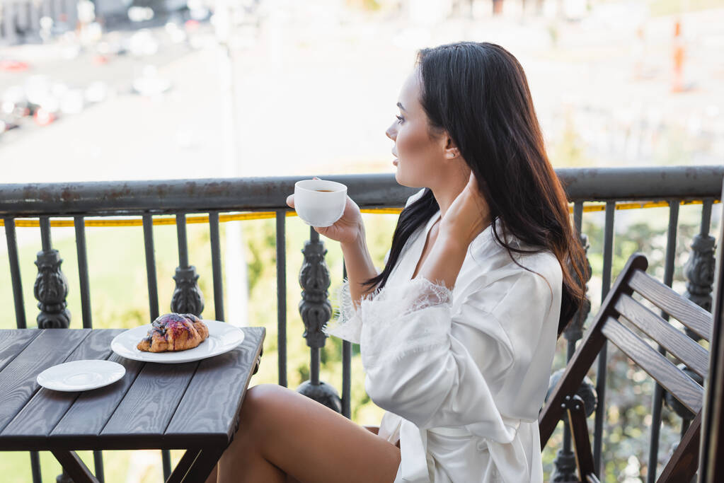 μελαχρινή γυναίκα σε λευκό ρόμπα πίνοντας τσάι και κάθεται στο μπαλκόνι - Φωτογραφία, εικόνα