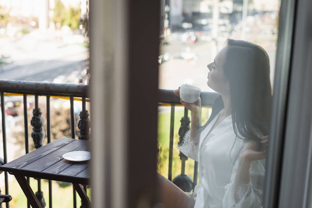 donna bruna con gli occhi chiusi che beve tè e si siede sul balcone - Foto, immagini