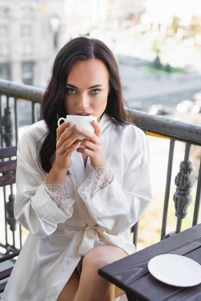 morena mujer en túnica blanca bebiendo té y sentado en el balcón - Foto, Imagen