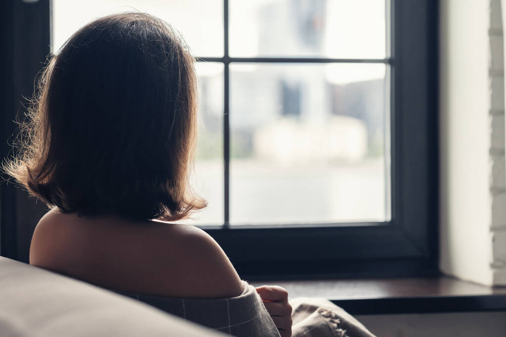 Vista trasera de la solitaria mujer joven aucasiática envuelta en un cuadros disfrutando sentado cerca de la ventana de los apartamentos.  - Foto, Imagen