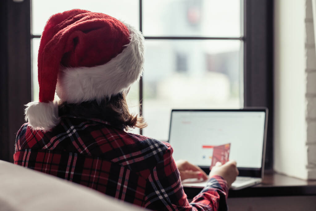 Nainen punaisessa joulupukin hatussa, jolla on luottokortti, tekee tilauksen ikkunan lähellä. Joulu verkkokaupoissa. Käsite - Valokuva, kuva