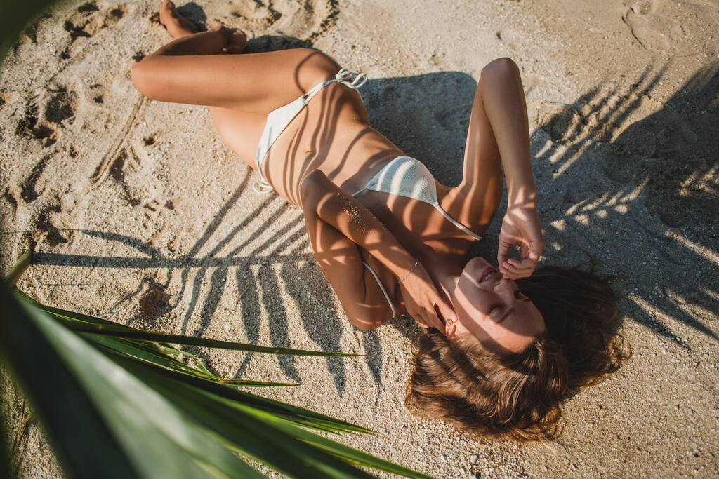 Genç seksi bir kadın kumsalda palmiye yaprağının altında yatıyor, vücudunda sanatsal gölgeler, beyaz bikini mayo, bronzlaşmış ten, ince vücut. - Fotoğraf, Görsel