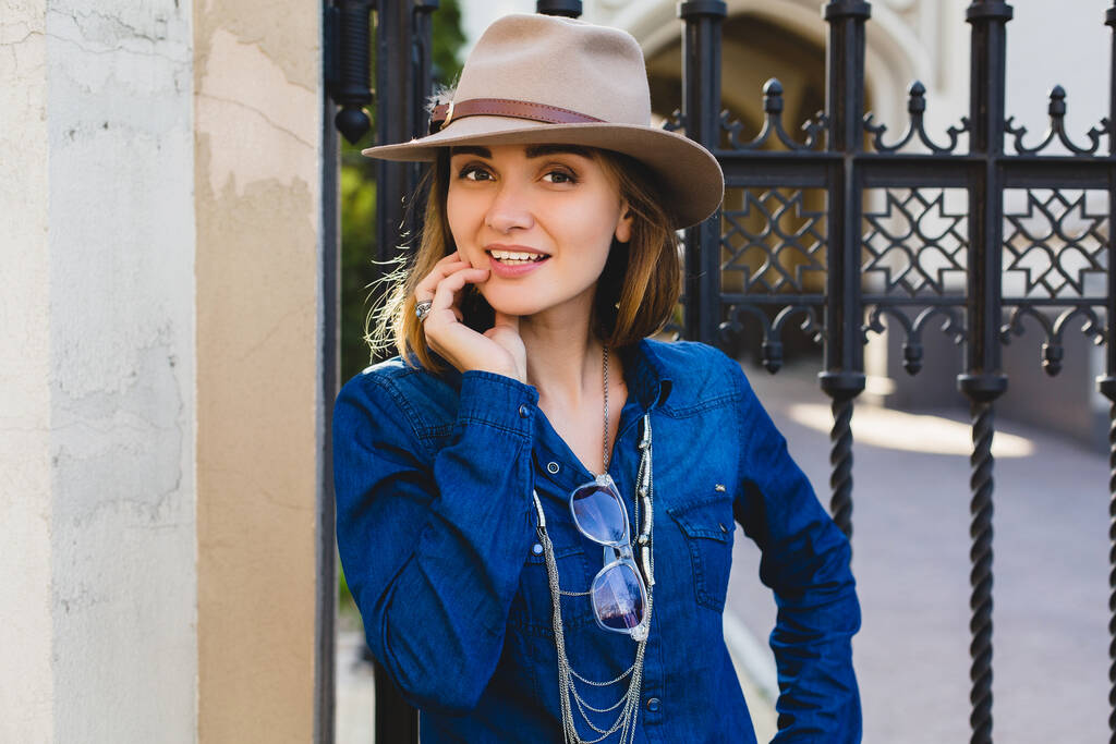 young stylish pretty happy woman smiling and posing, dressed in denim shirt, high heel shoes, hat and backpack, sunny day, good weather, city street, cool accessories, sunglasses, vacation - Photo, Image