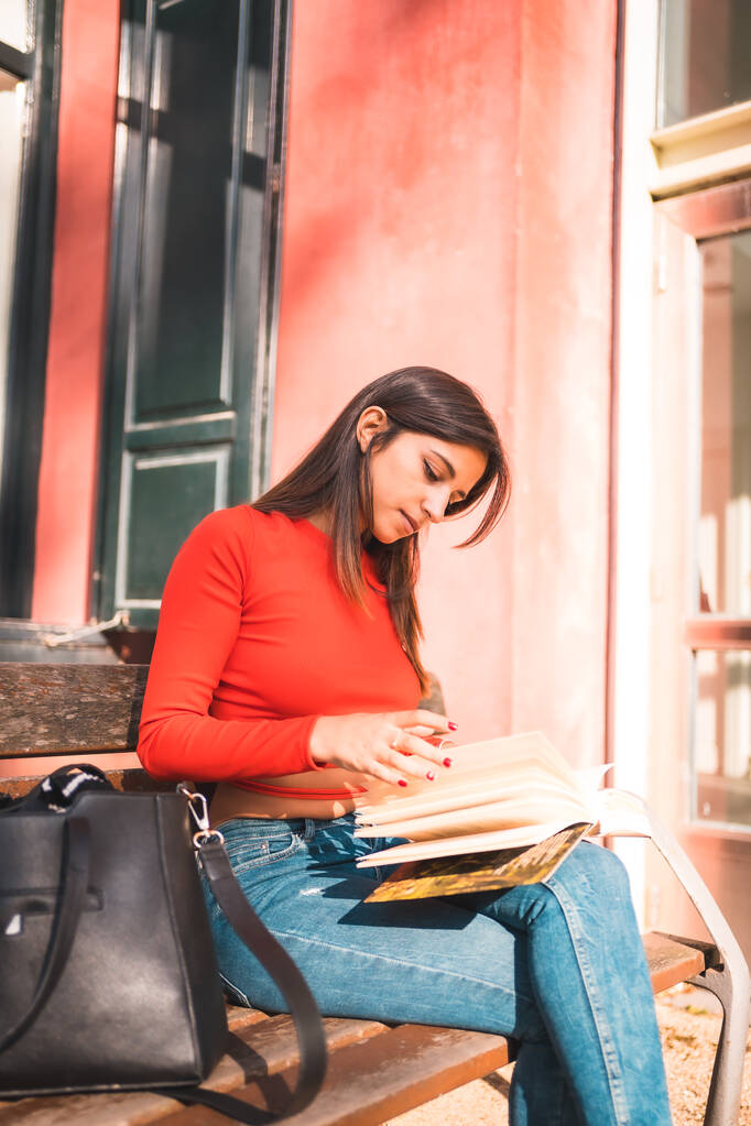 Estilo de vida. Jovem morena caucasiana com uma camisa vermelha sentada em um banco no parque lendo um livro. Foto vertical - Foto, Imagem