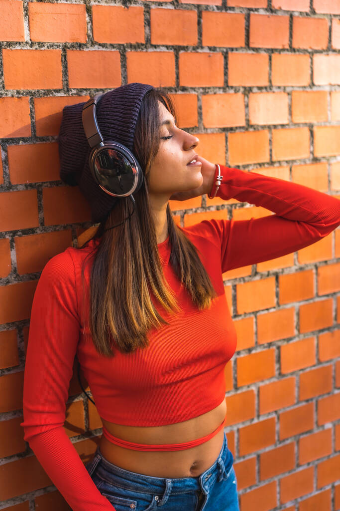 Styl życia, młoda brunetka z wełnianą czapką i czerwonym swetrem słuchającym muzyki opartej o ceglaną ścianę - Zdjęcie, obraz