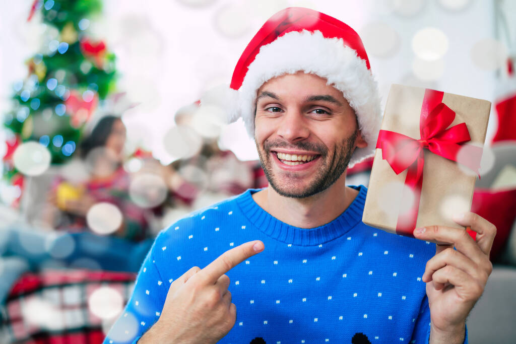 Junge aufgeregte gutaussehende Mann mit Weihnachtsmütze mit einem Weihnachtsgeschenk posiert und schaut in die Kamera auf einem Hintergrund von glücklichen Freunden zu Hause  - Foto, Bild