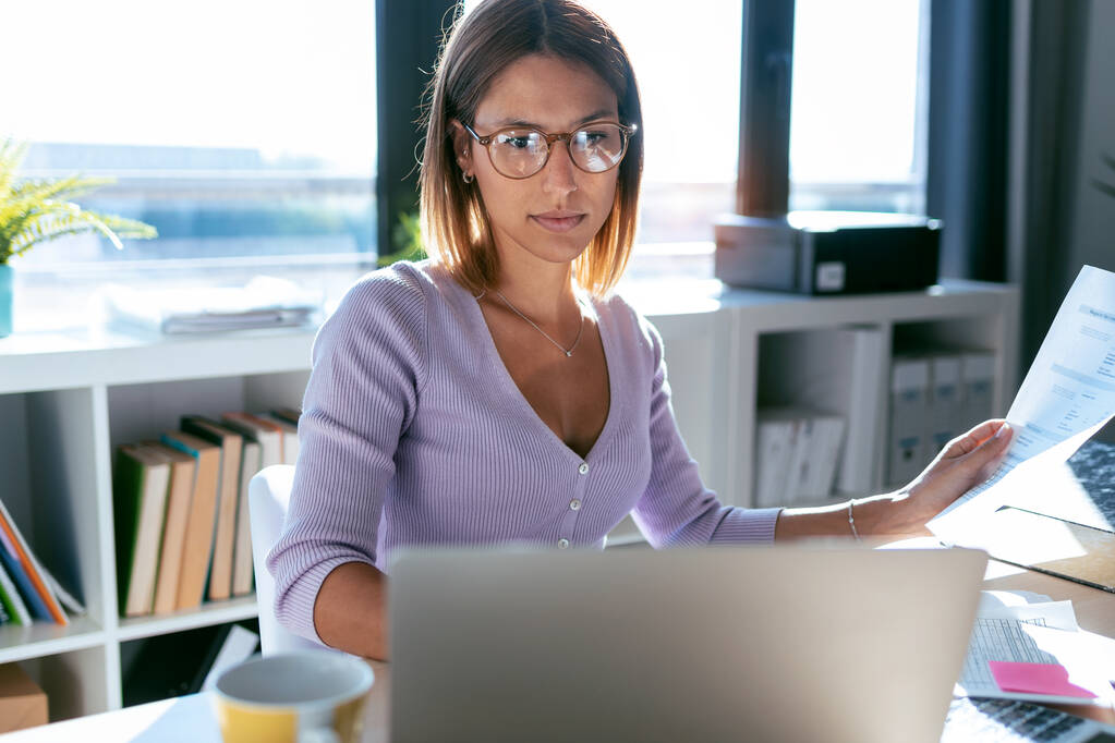 Aufnahme einer konzentrierten jungen Geschäftsfrau, die mit dem Computer arbeitet, während sie zu Hause im Büro einige Dokumente konsultiert. - Foto, Bild