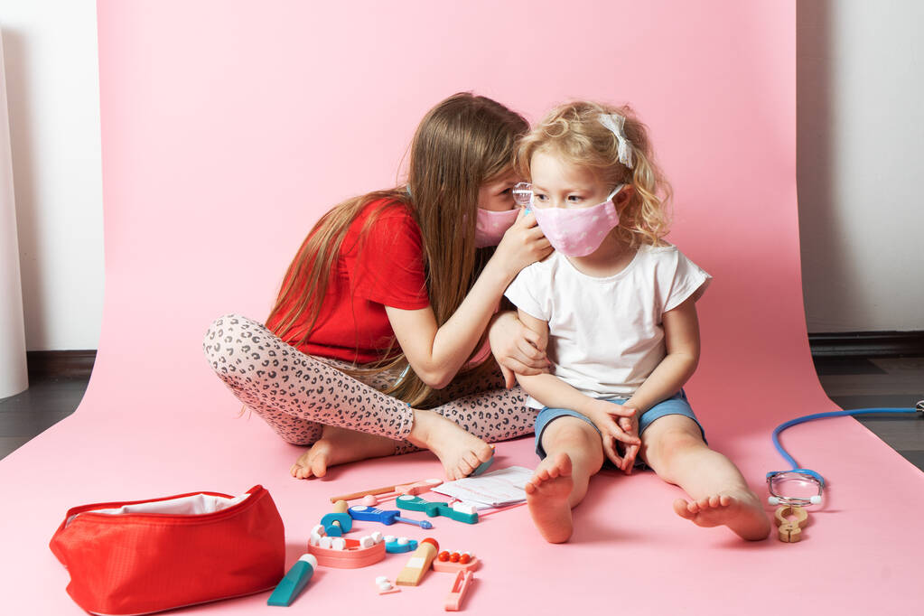 Osobní ochranné prostředky: dvě dívky v lékařských maskách a rukavicích si hrají na lékaře a používají lékařskou soupravu z hraček. - Fotografie, Obrázek