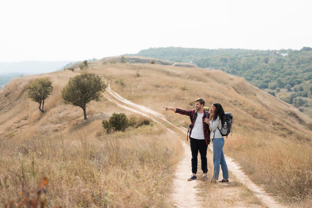 Smiling man with backpack pointing with finger near excited african american girlfriend on path during trip  - Photo, Image