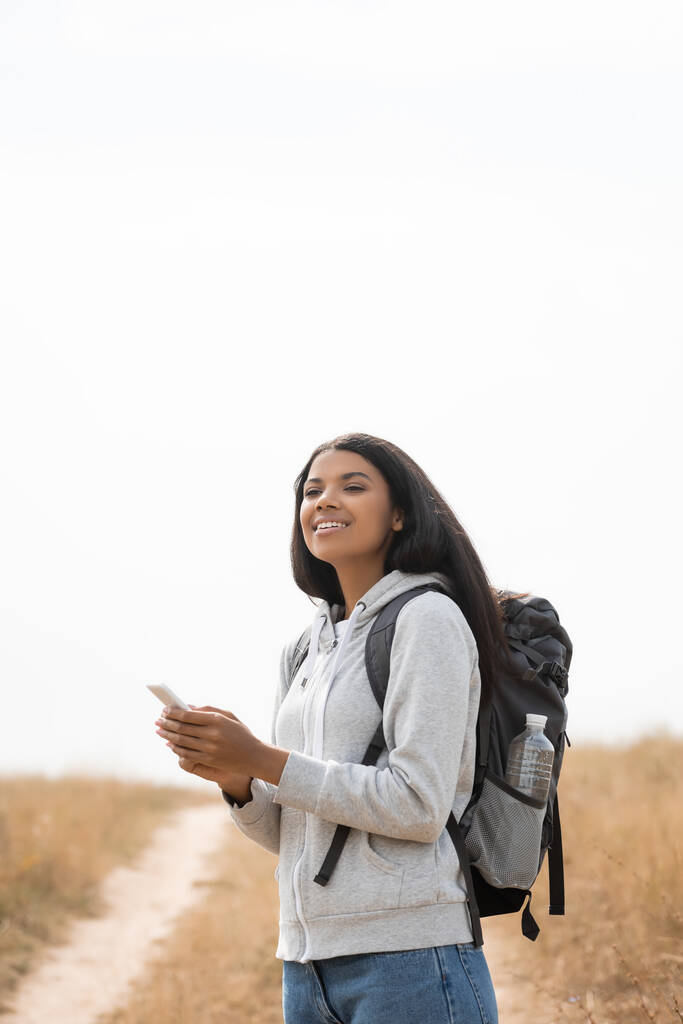 アフリカ系アメリカ人女性を笑顔でバックパックを持ってスマートフォンを持ってパス上のぼやけた背景  - 写真・画像