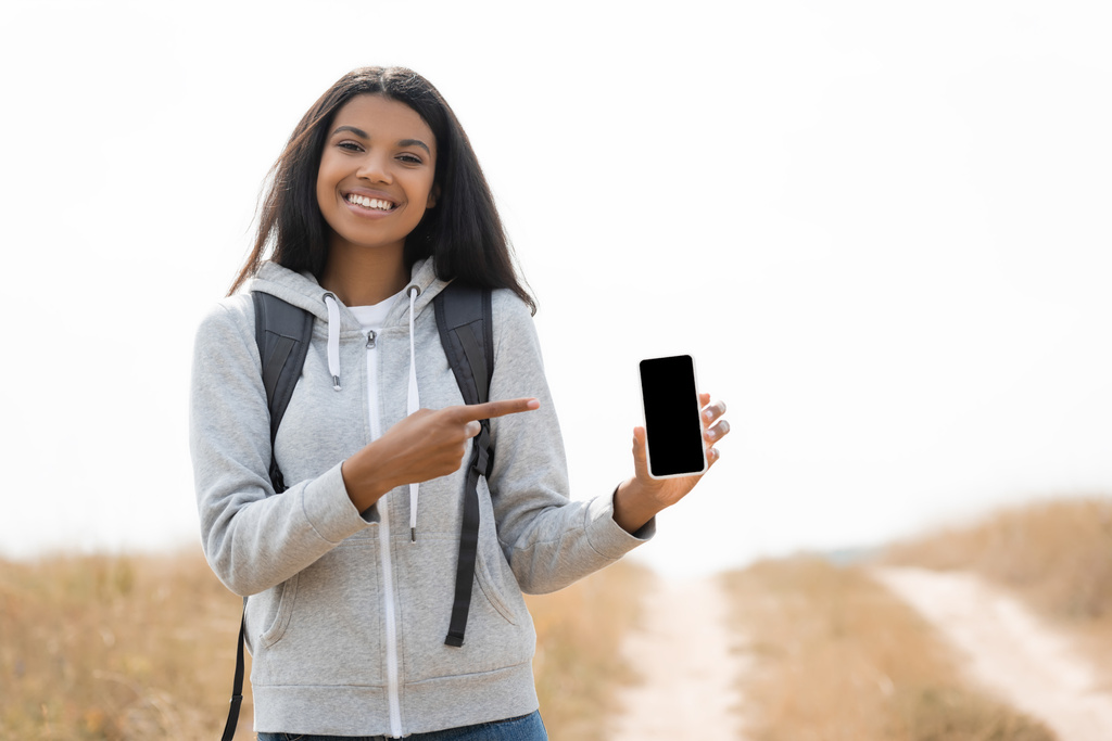 アフリカ系アメリカ人の旅行者が笑顔で指差す指でスマートフォンの近くに空白の画面でパス上のぼやけた背景  - 写真・画像