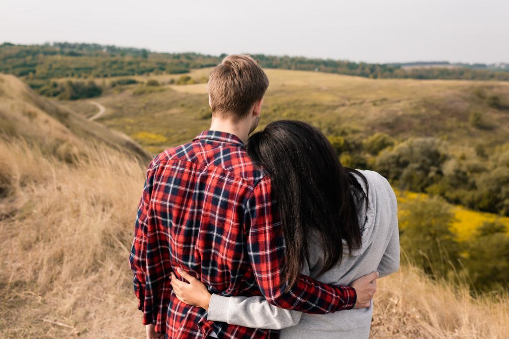 Vue arrière d'un couple multiethnique embrassant un paysage herbeux sur fond flou - Photo, image