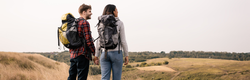Usmívající se mezirasový pár s batohy drží ruce s travnatými kopci na rozmazaném pozadí, banner  - Fotografie, Obrázek