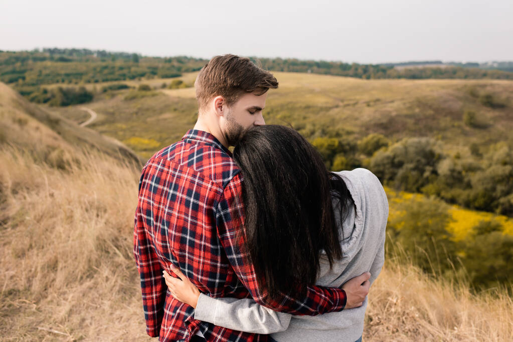 Achteraanzicht van jonge man knuffelen Afrikaans amerikaanse vriendin met gras heuvels op wazig achtergrond  - Foto, afbeelding