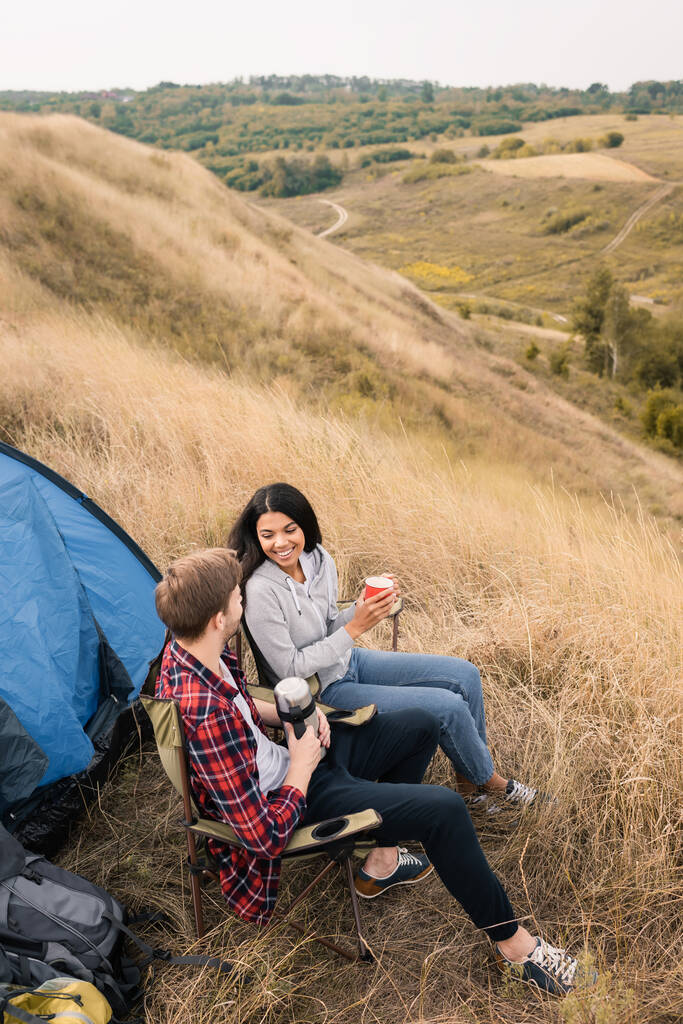 Couple interracial souriant tenant tasse et thermos près de la tente pendant le camping dans le champ   - Photo, image