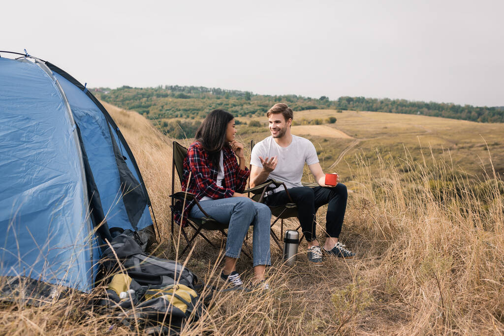 Smiling man with cup talking to african american girlfriend near backpacks and tent on meadow  - Photo, Image