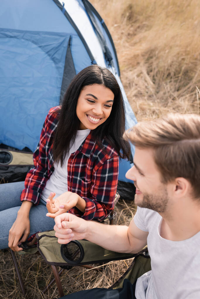 Lächelnde Afroamerikanerin sieht Freund auf verschwommenem Vordergrund in der Nähe von Zelt beim Zelten an  - Foto, Bild
