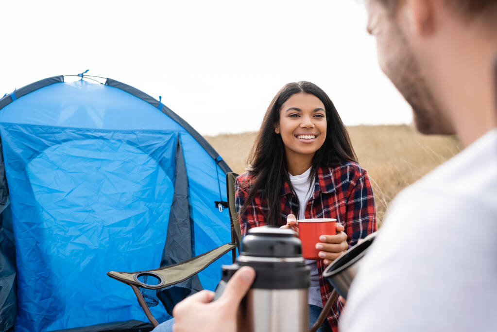 Веселая африканская американка держит чашку возле бойфренда с термосом на размытом переднем плане и палаткой на открытом воздухе  - Фото, изображение