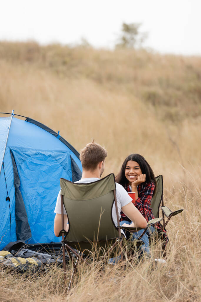 Sorrindo Africano americano mulher segurando copo e olhando para namorado perto da tenda no gramado gramado  - Foto, Imagem