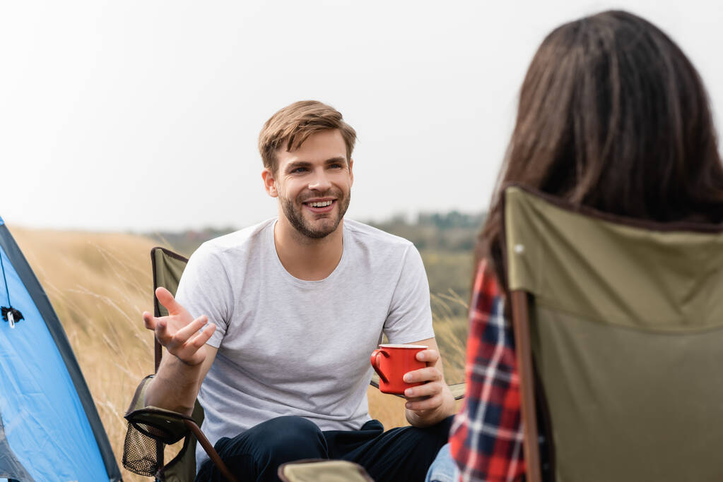 Улыбающийся мужчина с чашкой разговаривает с девушкой на размытом переднем плане во время похода  - Фото, изображение
