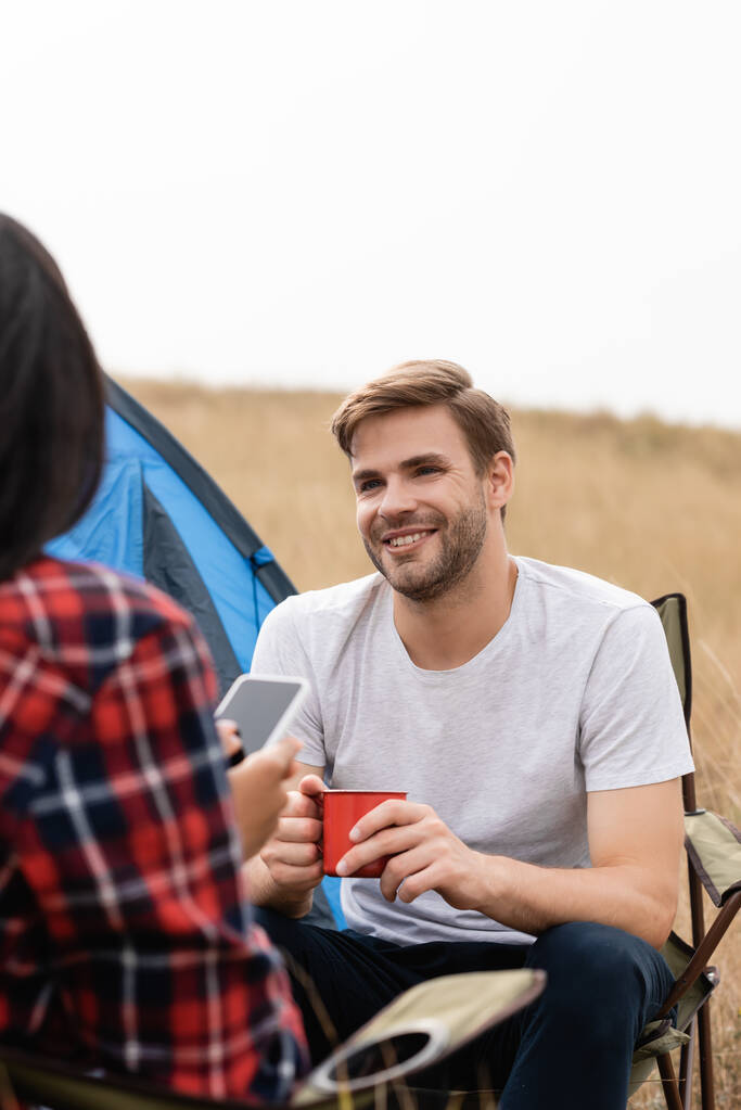 Hymyilevä mies tilalla cup lähellä afrikkalainen amerikkalainen tyttöystävä älypuhelin hämärtynyt etualalla aikana camping  - Valokuva, kuva
