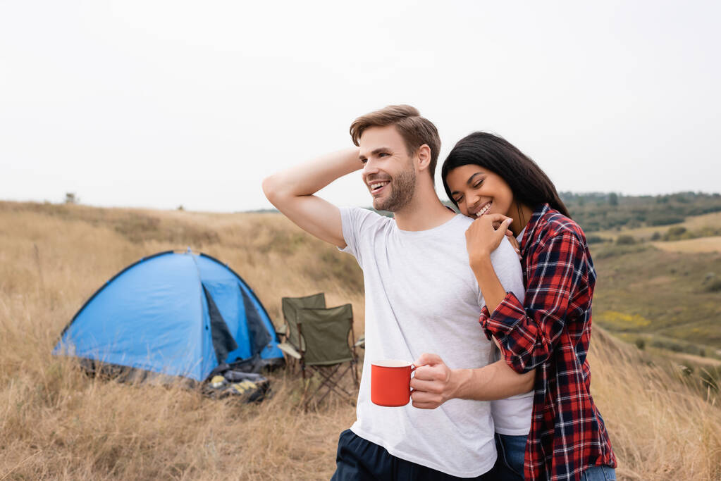 Femme afro-américaine avec les yeux fermés étreignant petit ami avec tasse près de la tente au premier plan flou  - Photo, image