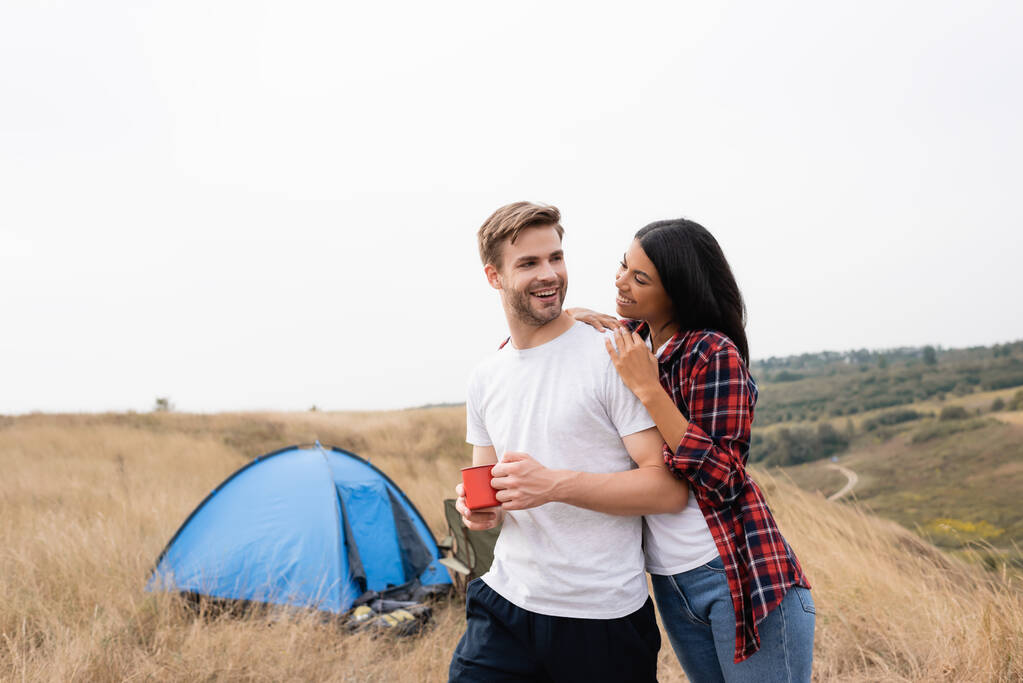 Sourire femme afro-américaine étreignant petit ami avec tasse près de la tente sur fond flou à l'extérieur  - Photo, image