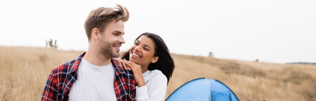 Smiling man looking at african american girlfriend with tent and field on blurred background, banner  - Photo, Image