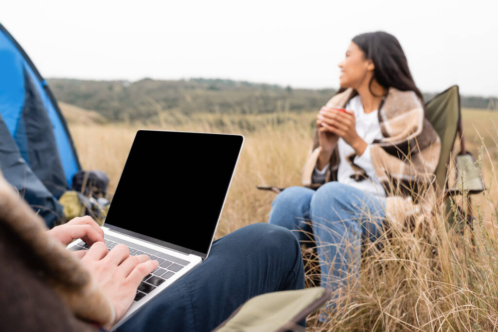 Uomo utilizzando laptop vicino fidanzata afro-americana con coppa su sfondo sfocato durante il campeggio in campo  - Foto, immagini