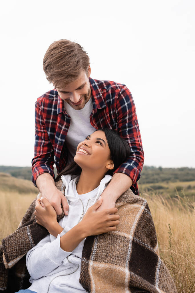 Smiling man embracing african american girlfriend wrapped in blanket on meadow  - Photo, Image