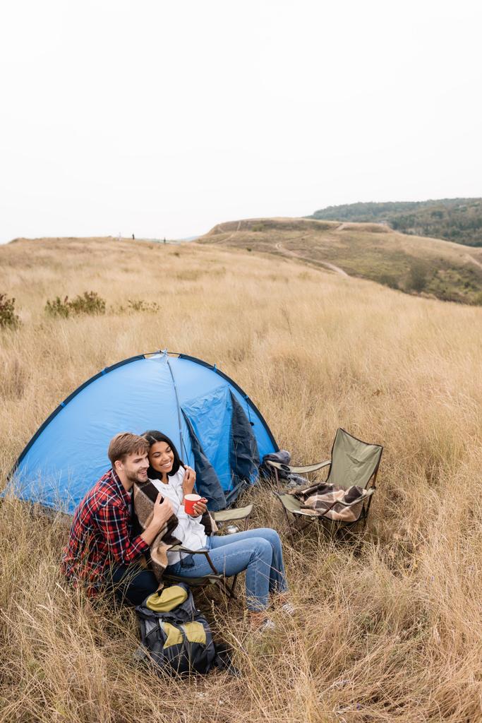 Улыбающаяся межрасовая пара с одеялом и чашкой, сидящая возле палатки на травянистой лужайке  - Фото, изображение