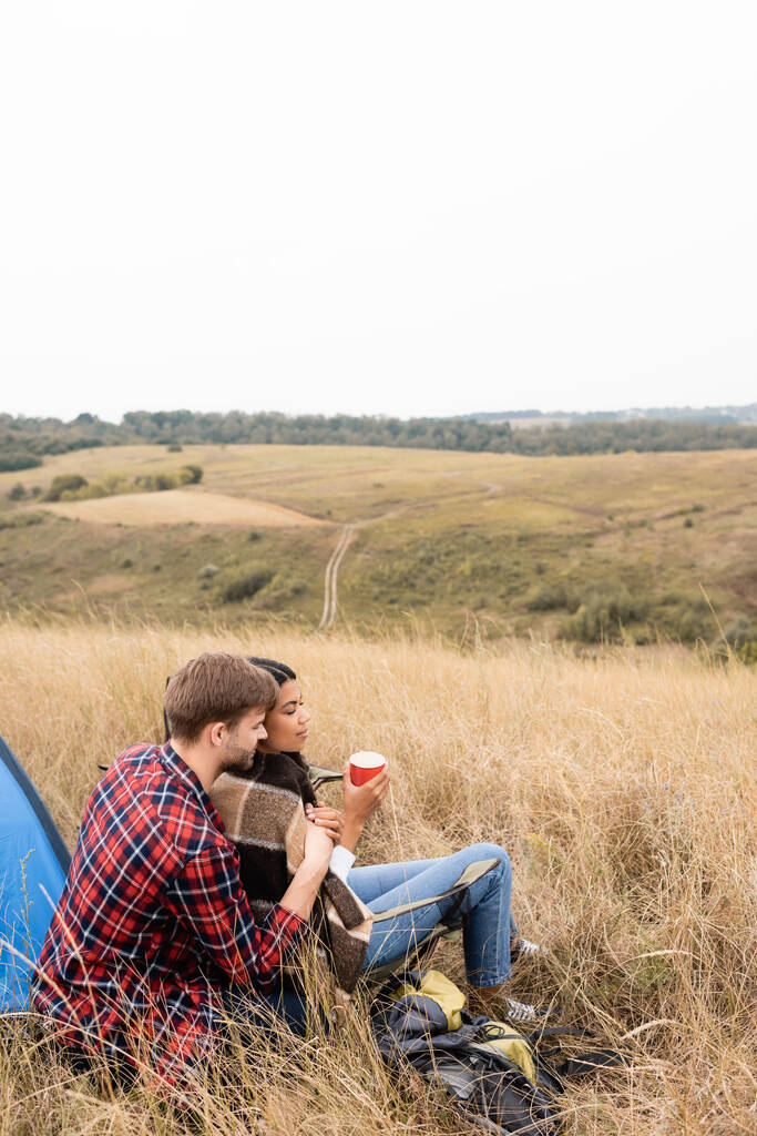 Homme étreignant petite amie afro-américaine avec tasse pendant le camping sur un terrain herbeux  - Photo, image