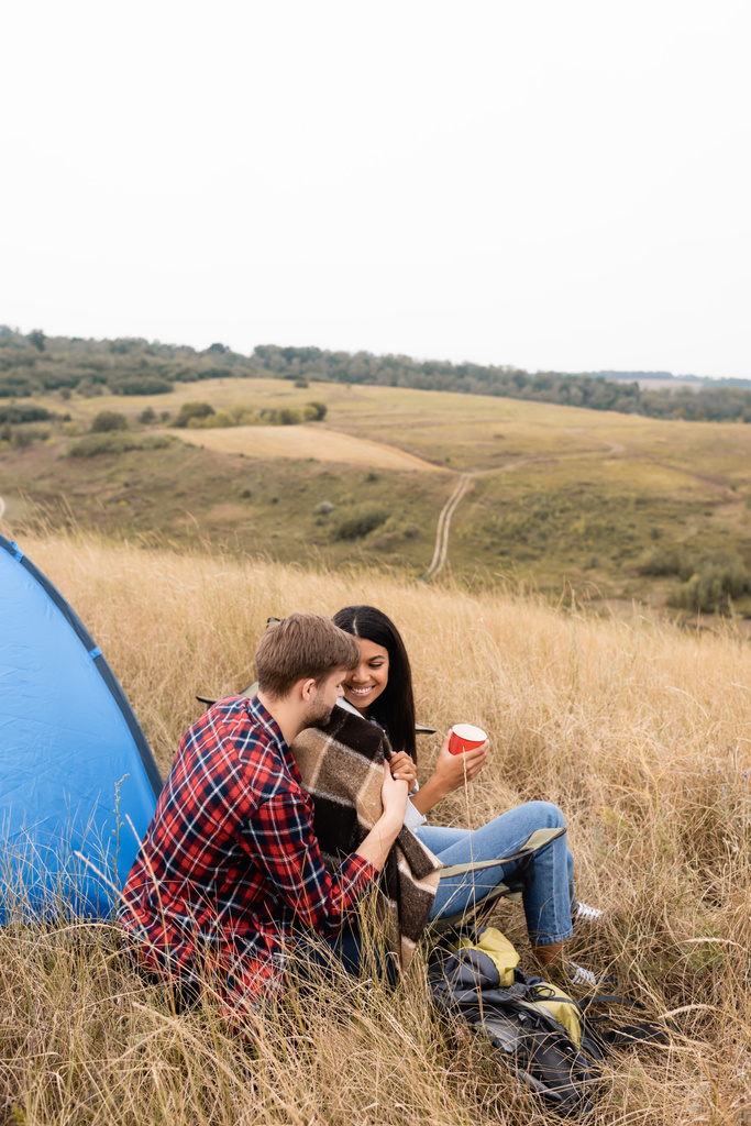 Multiethnic couple with blanket and cup sitting near backpack and tent on field  - Photo, Image