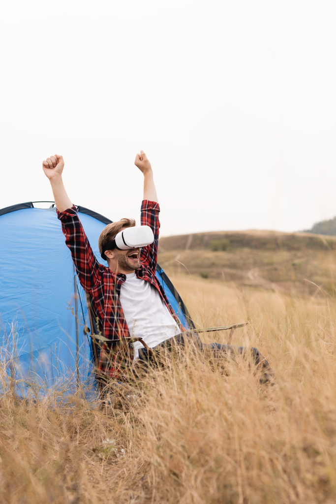 Opgewonden man toont ja gebaar tijdens het gebruik van vr headset in de buurt van tent tijdens het kamperen  - Foto, afbeelding