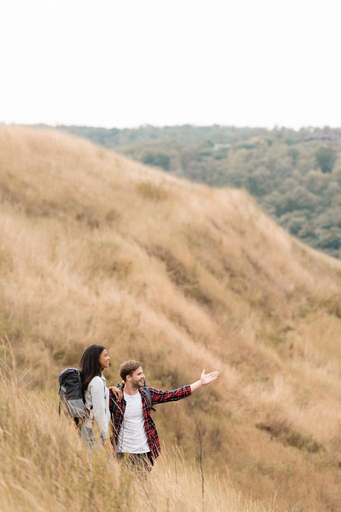 Mosolygós férfi mutató kézzel, miközben áll közel afro-amerikai barátnő hátizsák a dombon  - Fotó, kép