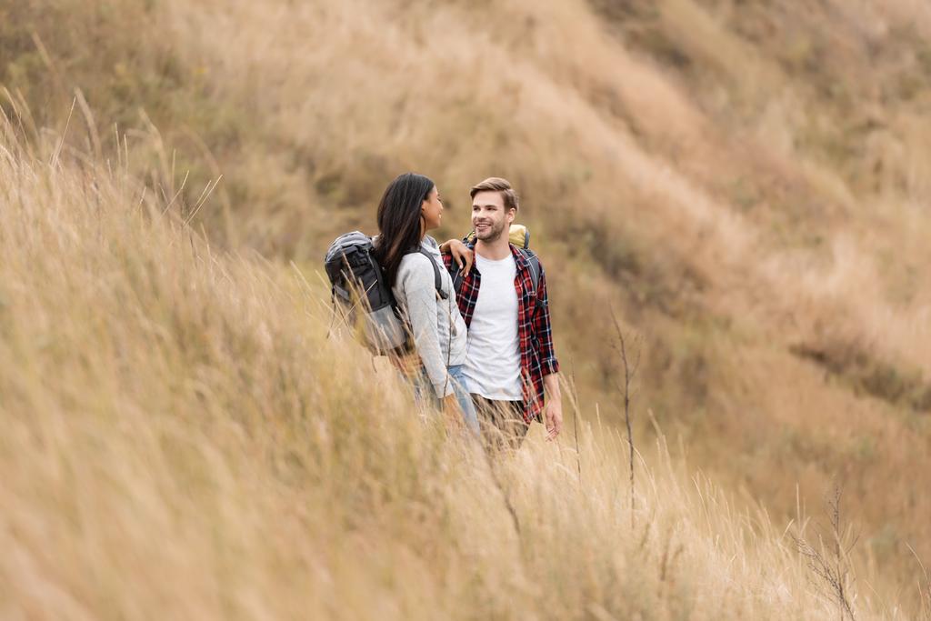 Usmívající se mezirasový pár s batohy při pohledu na sebe na plynatém kopci  - Fotografie, Obrázek