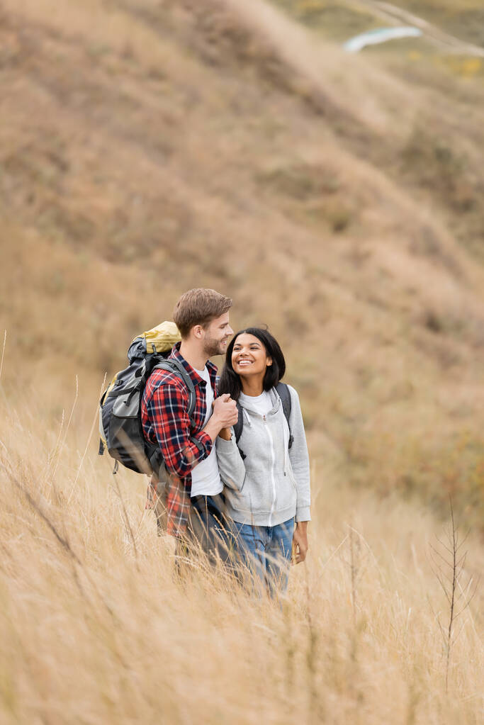 Usmívající se multietnický pár s batohy držící se za ruce na travnatém kopci  - Fotografie, Obrázek