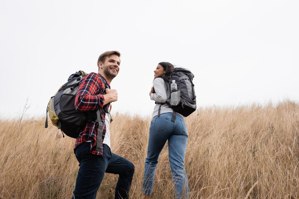 Giovane escursionista sorridente vicino alla ragazza afro-americana che cammina sulla collina erbosa  - Foto, immagini