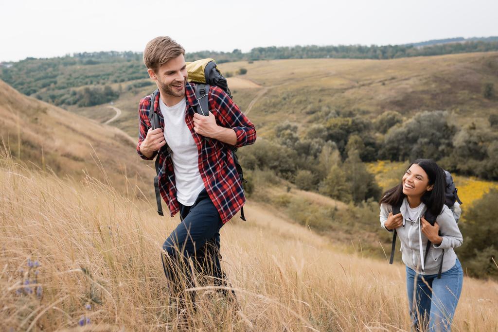Homme souriant avec sac à dos marchant près de petite amie afro-américaine sur la colline avec de l'herbe  - Photo, image