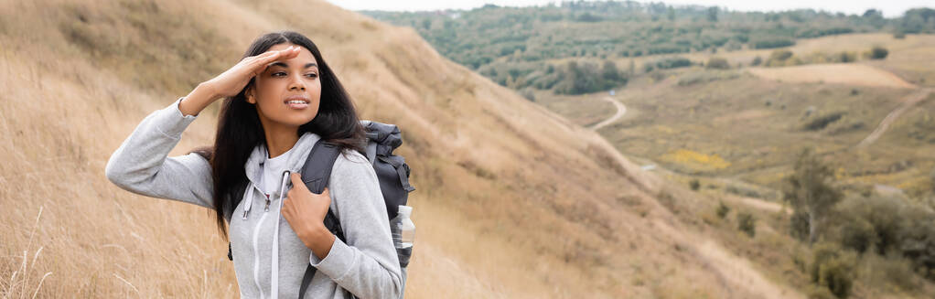 Mosolygó afro-amerikai túrázó néz félre, miközben áll a dombon utazás közben, banner  - Fotó, kép