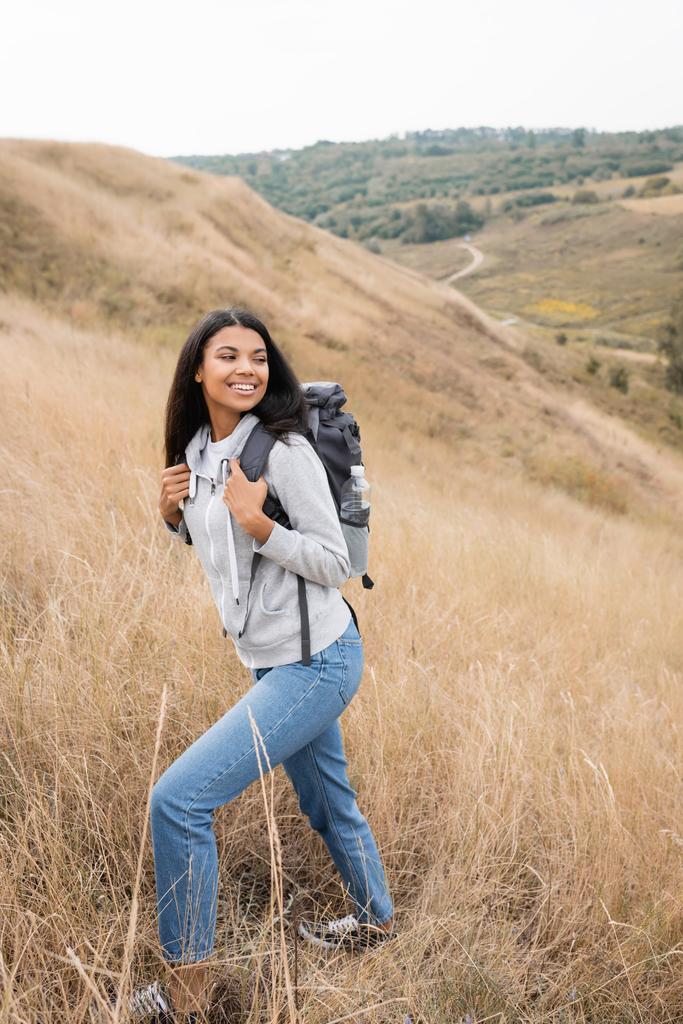 Pozitív afro-amerikai túrázó kezében hátizsák, miközben séta a dombon fű - Fotó, kép