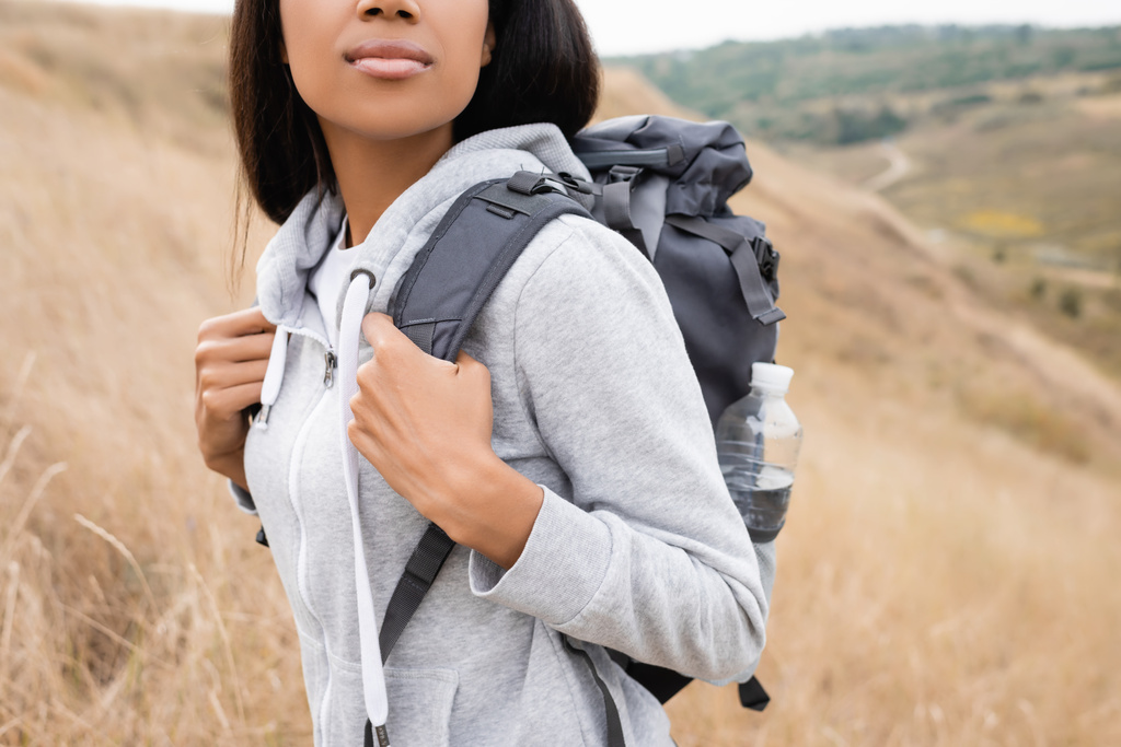 Vista recortada de mujer afroamericana con mochila de pie en la colina con paisaje borroso en el fondo  - Foto, Imagen