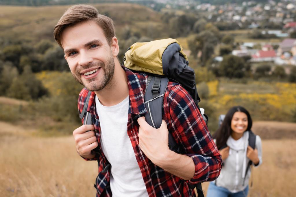Turista sorrindo para a câmera enquanto caminhava perto de mulher americana africana no fundo borrado ao ar livre  - Foto, Imagem