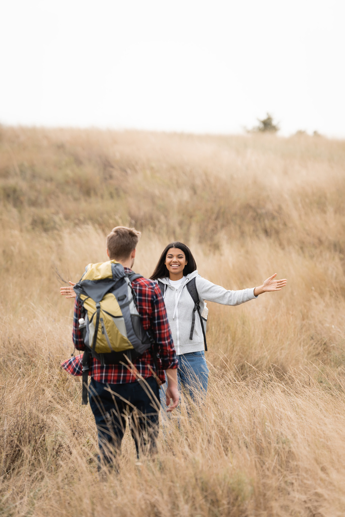 Smiling african american woman smiling at boyfriend with backpack on blurred foreground on grassy hill  - Photo, Image