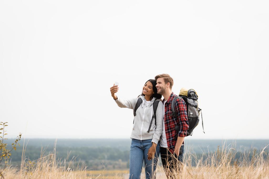 Hymyilevä rotujenvälinen pari reppuineen puhuu selfie älypuhelimella nurmikolla  - Valokuva, kuva